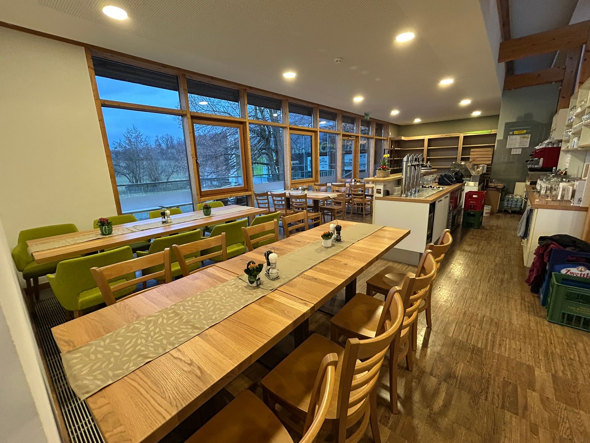 Ein gemütliches Café mit Holztischen und grünen Stühlen, großen Fenstern und einer Theke mit Zapfanlage.