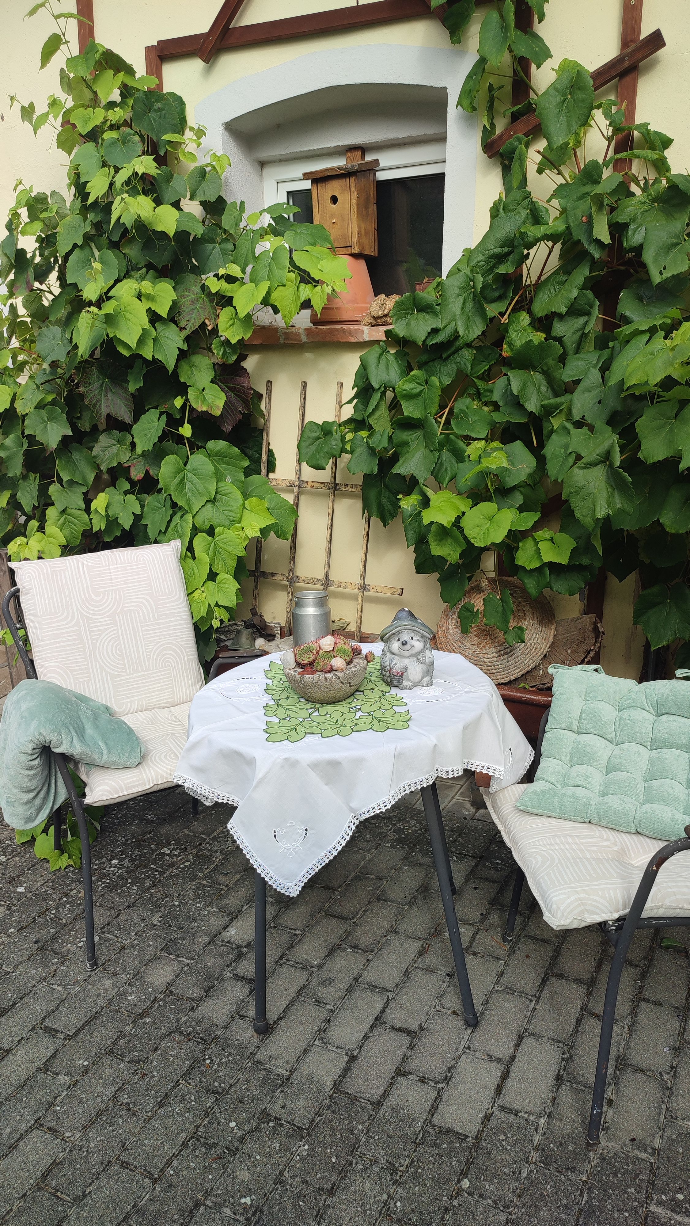 Ein gemütlicher Sitzbereich im Freien mit zwei Stühlen, einem Tisch mit Tischdecke, dekoriert mit einer Figur und Pflanzen im Hintergrund.