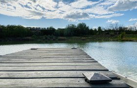 Holzsteg am Ziegelofenteich in Zellerndorf, umgeben von Bäumen und blauem Himmel mit Wolken.