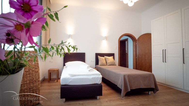 Ein gemütliches Zweibettzimmer mit Blumen im Vordergrund, zwei Betten, einem Schrank und einer Lampe an der Decke.