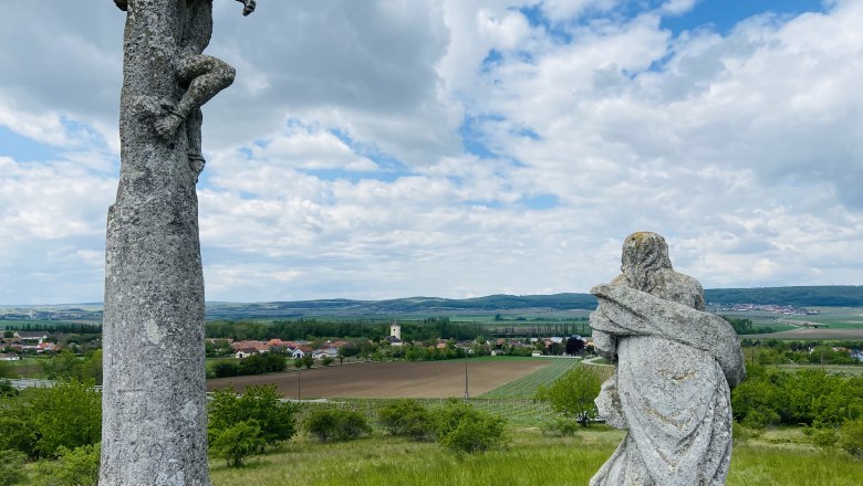 Zwei Steinskulpturen auf einem Hügel mit Blick auf eine ländliche Landschaft und ein Dorf im Hintergrund.