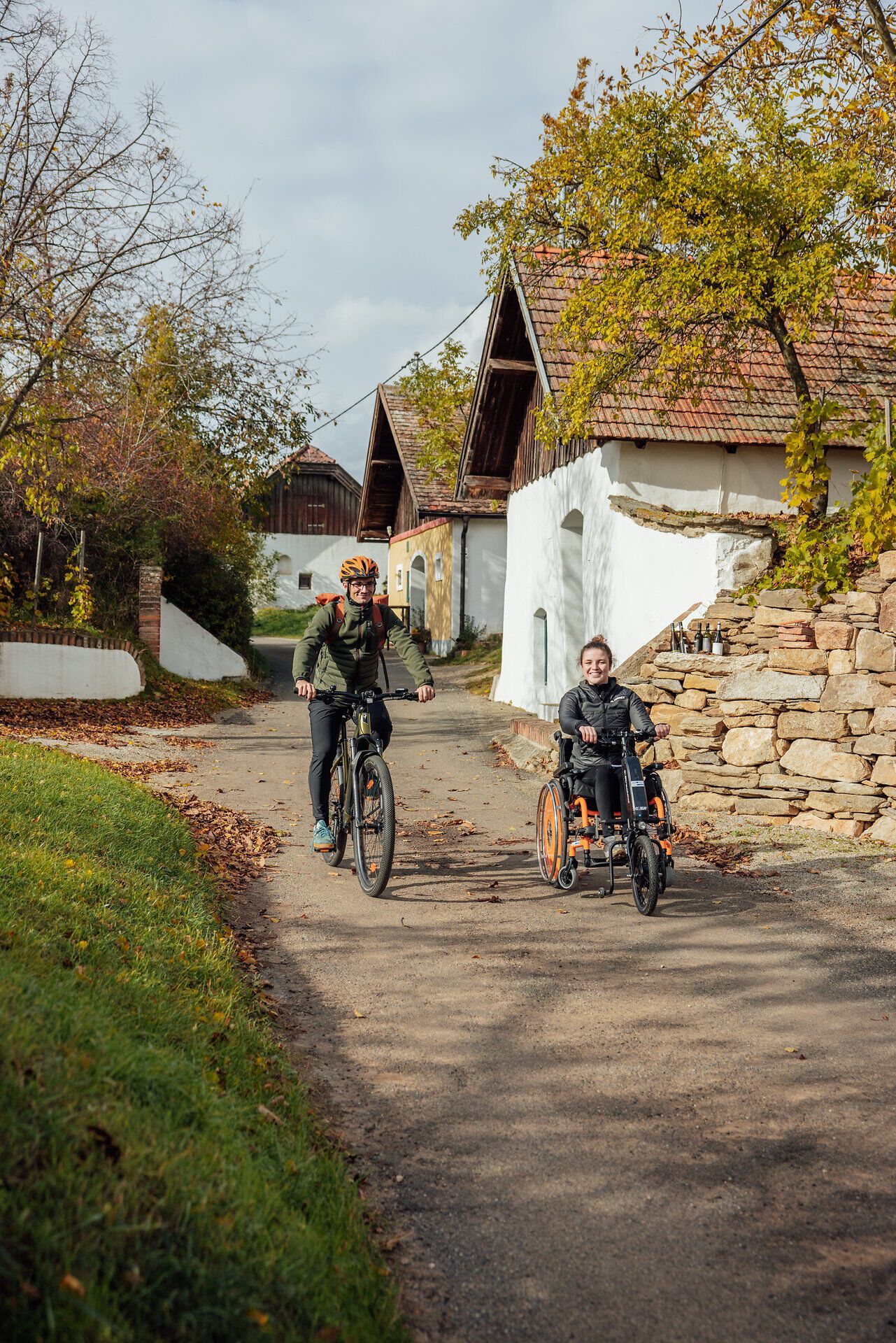 Eine Frau im E-Rollstuhl mit Zusatzrad bewegt sich neben einem Mann auf einem Fahrrad vorbei an Presshäusern durch die Öhlbergkellergasse in Pillersdorf.