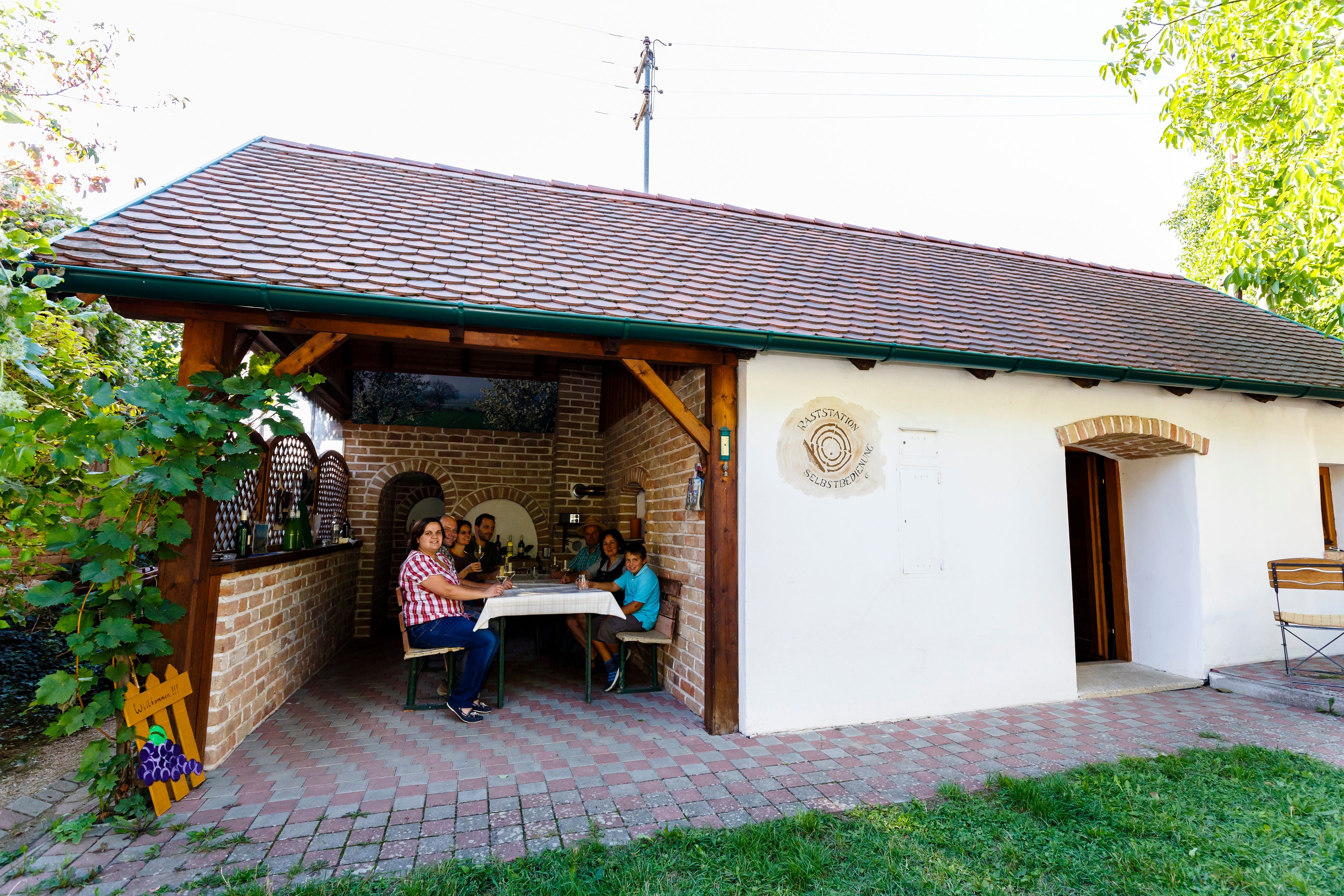 Eine Familie sitzt an einem Tisch in einem überdachten Außenbereich eines Gebäudes mit der Aufschrift 'Tankstation Selbstbedienung'.