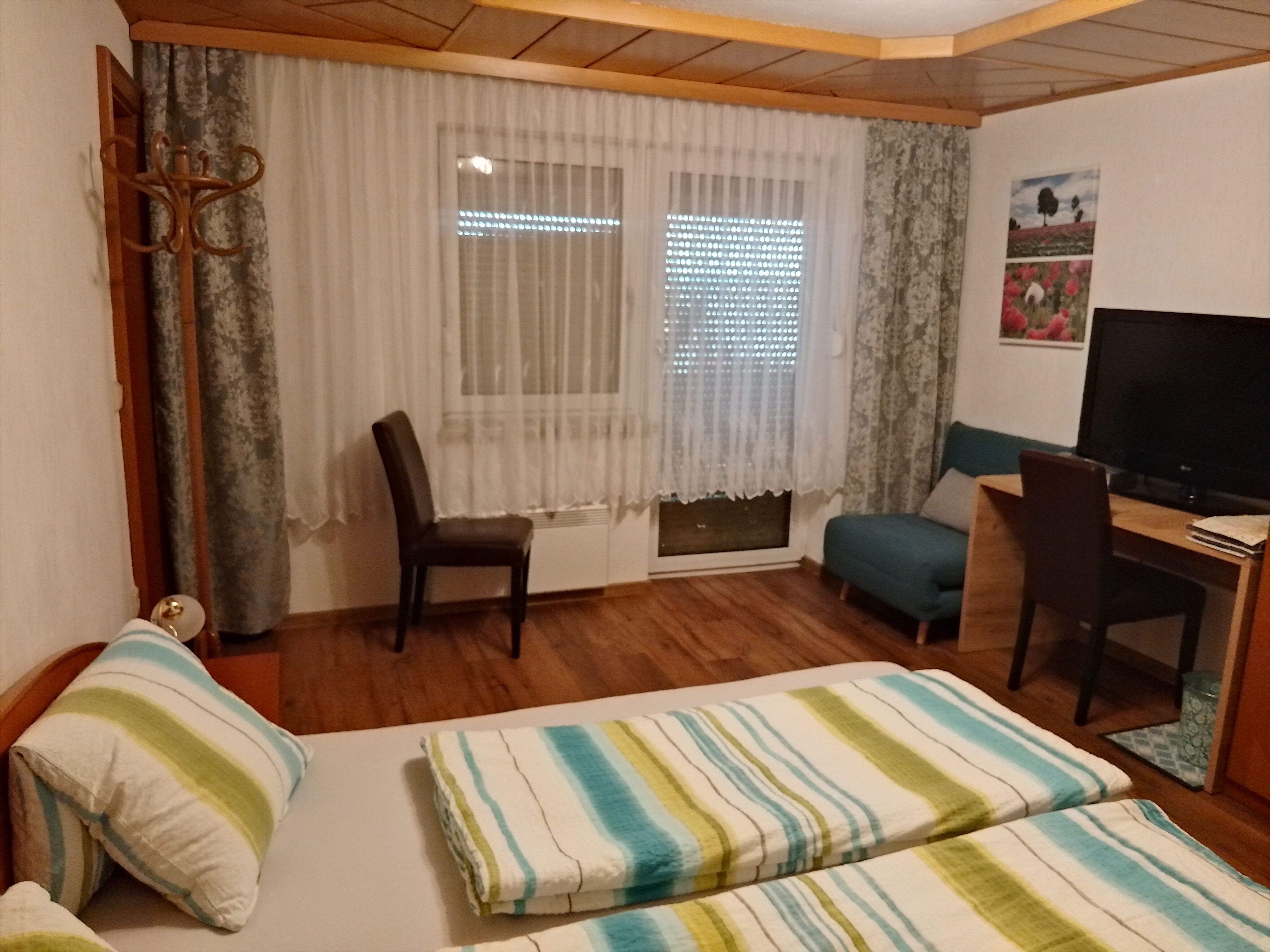 Ein gemütliches Privatzimmer mit Doppelbett, Schreibtisch, Stuhl und Fernseher. Vorhänge und ein Bild an der Wand ergänzen die Einrichtung.