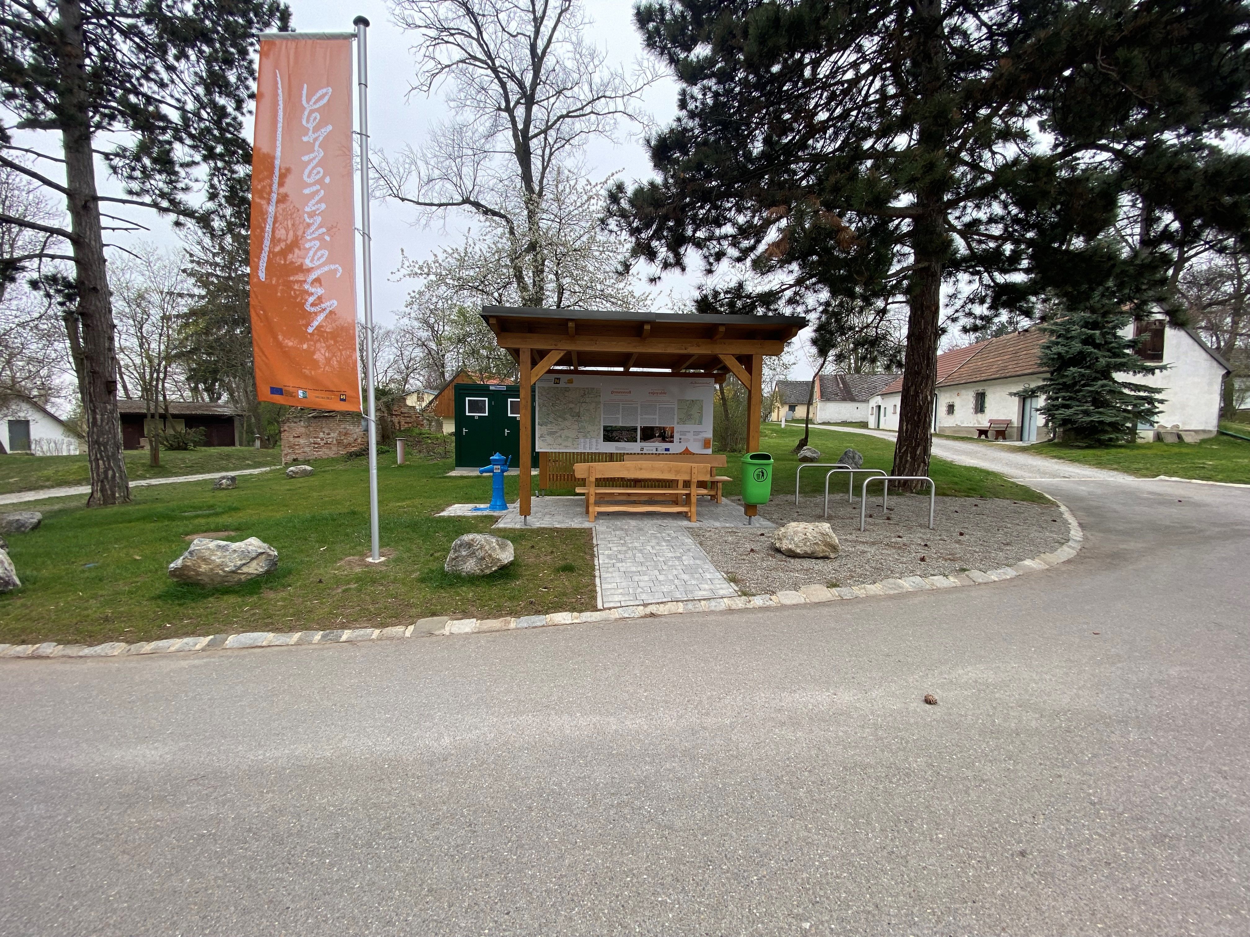 Ein Rastplatz mit überdachtem Sitzbereich, Infotafel und Fahrradständern. Daneben weht eine orange Fahne. Im Hintergrund sind Bäume und Gebäude zu sehen.