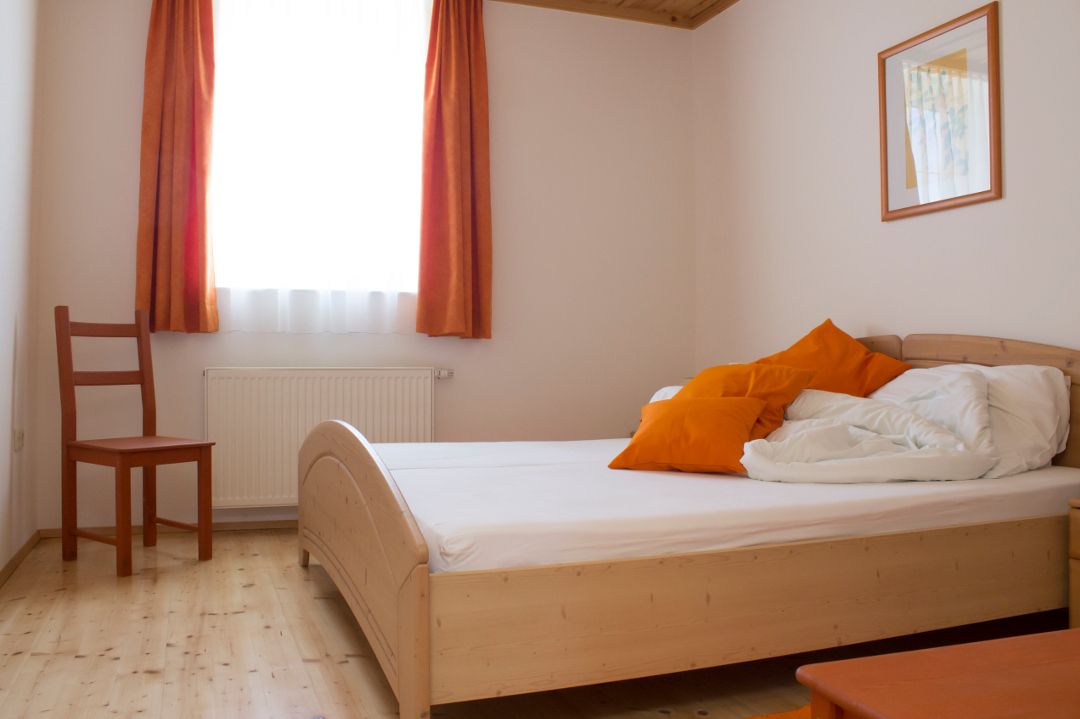 Ein einfach eingerichtetes Doppelzimmer mit Holzbett, orangefarbenen Kissen und Vorhängen, einem Stuhl und einem Bild an der Wand.