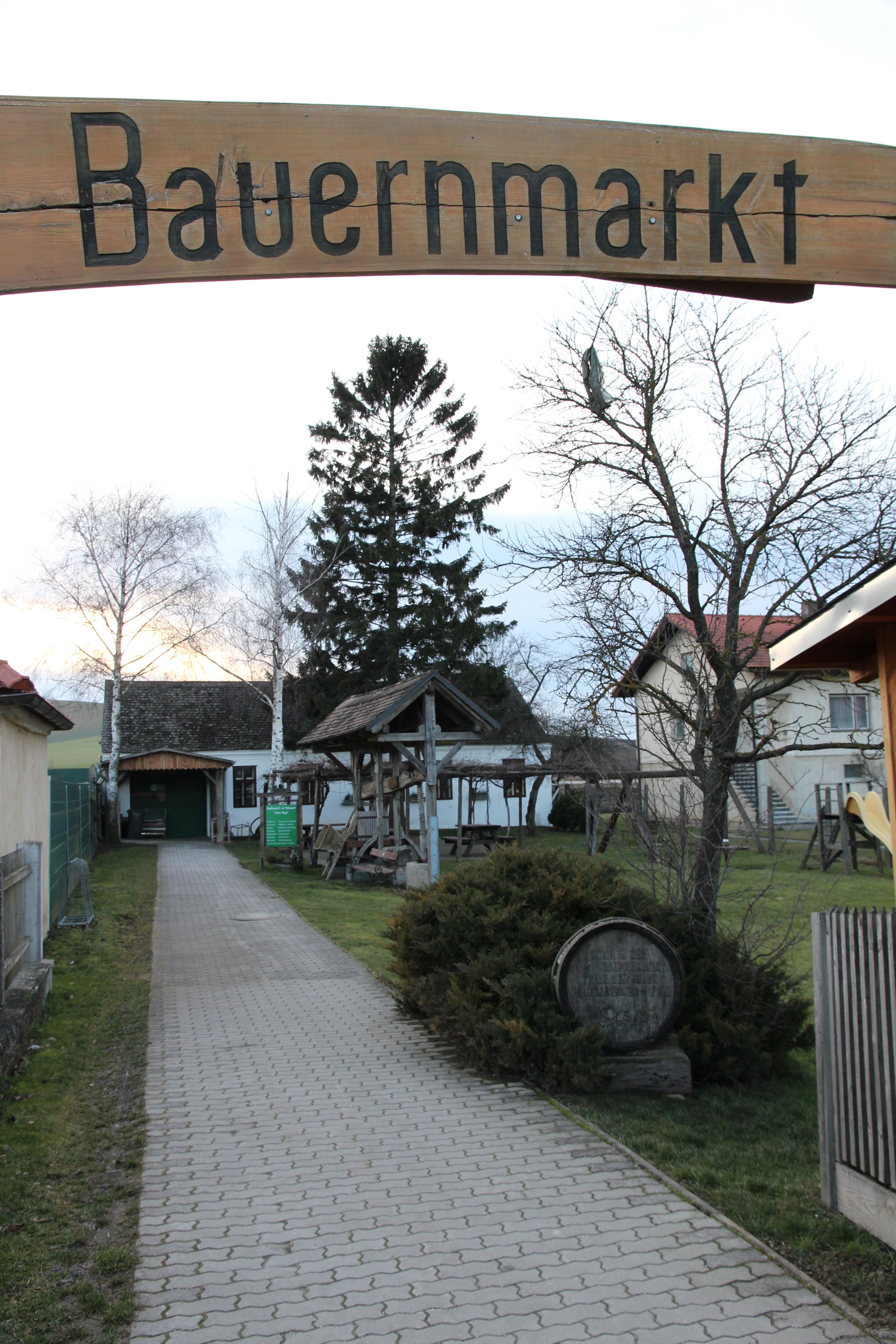 Eingang zu einem Bauernmarkt mit einem Holzschild und einem gepflasterten Weg, umgeben von Bäumen und Gebäuden.