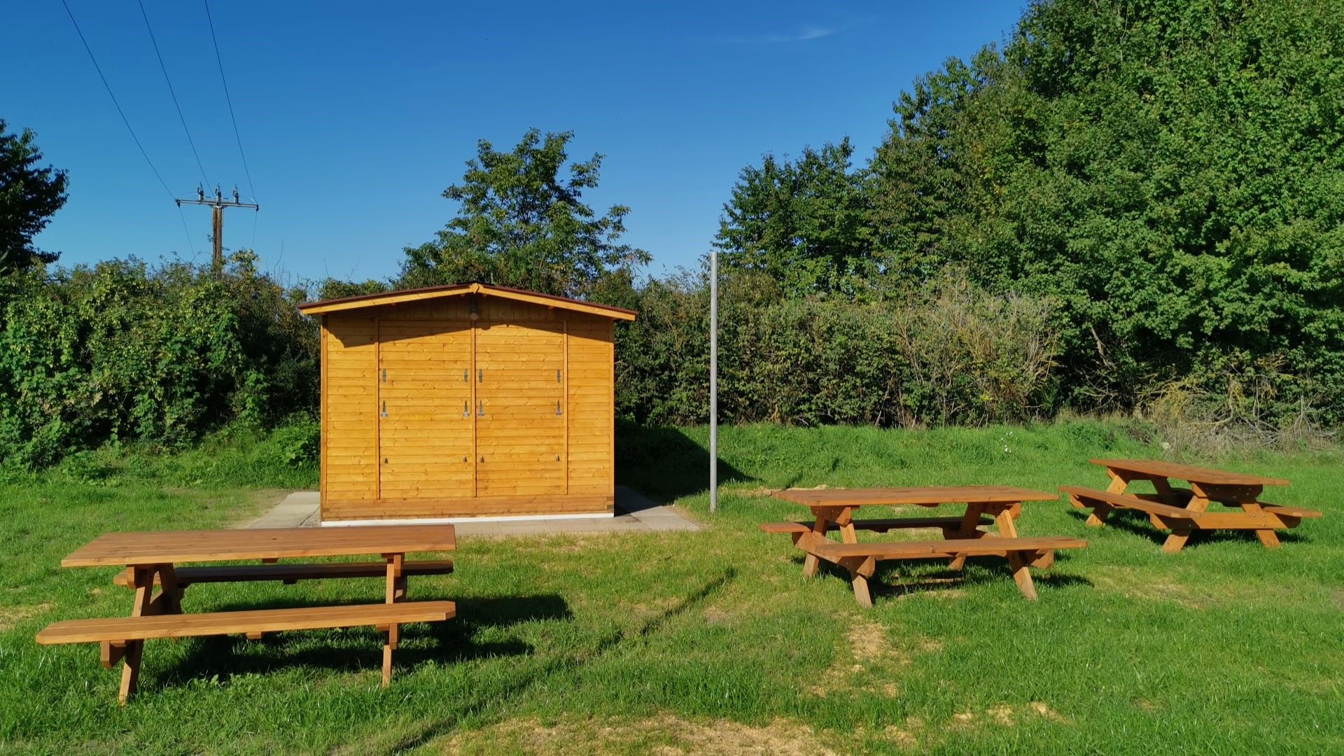 Wooden hut with picnic tables on a meadow, surrounded by trees and bushes under a blue sky.