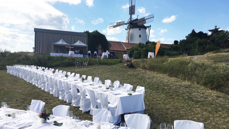 Lange Tische im Freien mit weißen Tischdecken, im Hintergrund eine Windmühle.