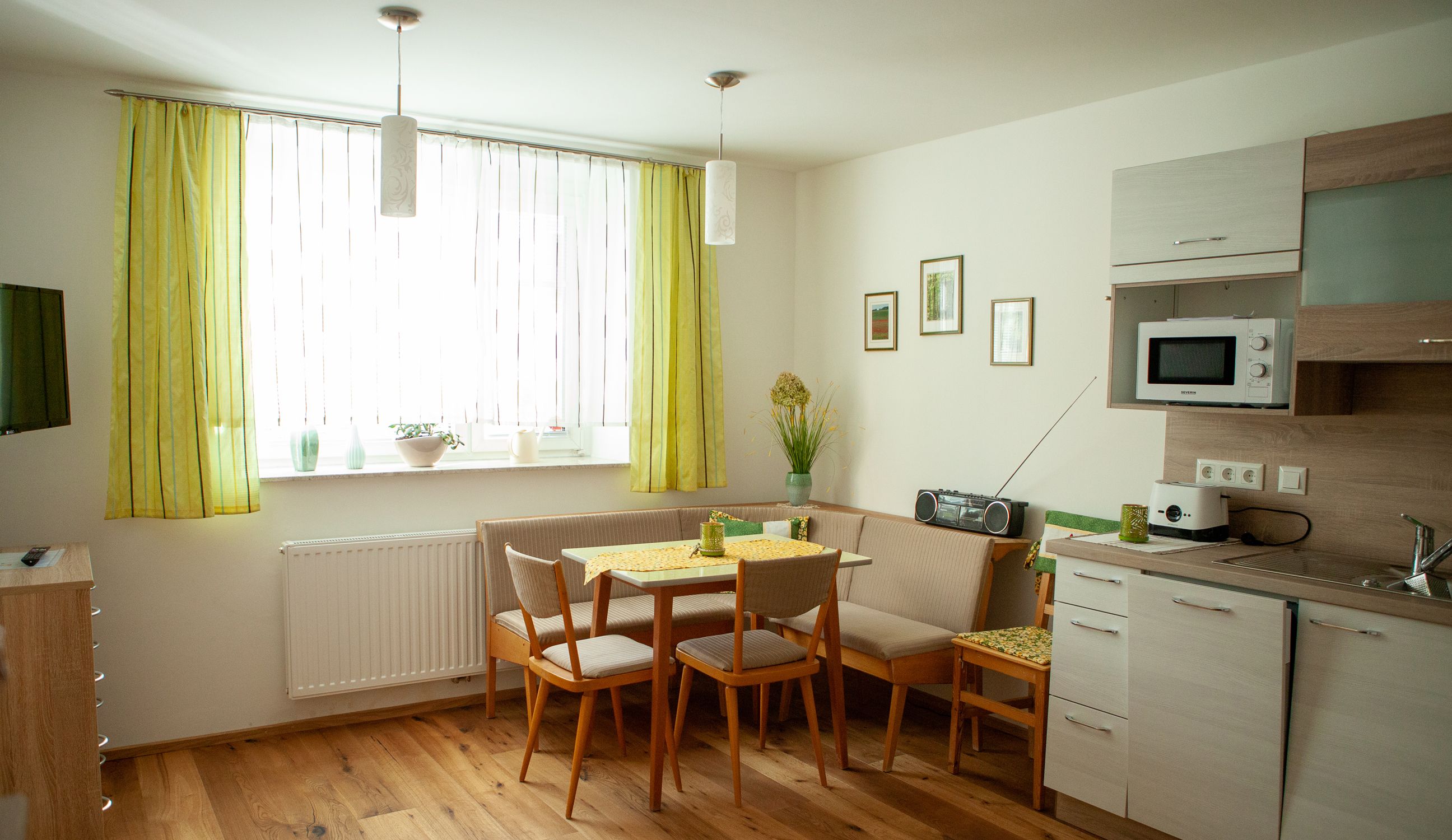 Gemütliche Wohnküche mit Esstisch, Eckbank, Küchenzeile und grünen Vorhängen.