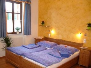 Ein gemütliches Doppelzimmer mit Holzmöbeln, blauer Bettwäsche und gelber Wand. Pflanzen und Fenster mit blauen Vorhängen sind sichtbar.