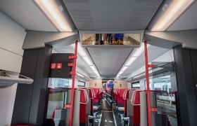 Innenansicht eines modernen Zugabteils mit roten und grauen Sitzen und einer digitalen Anzeigetafel.