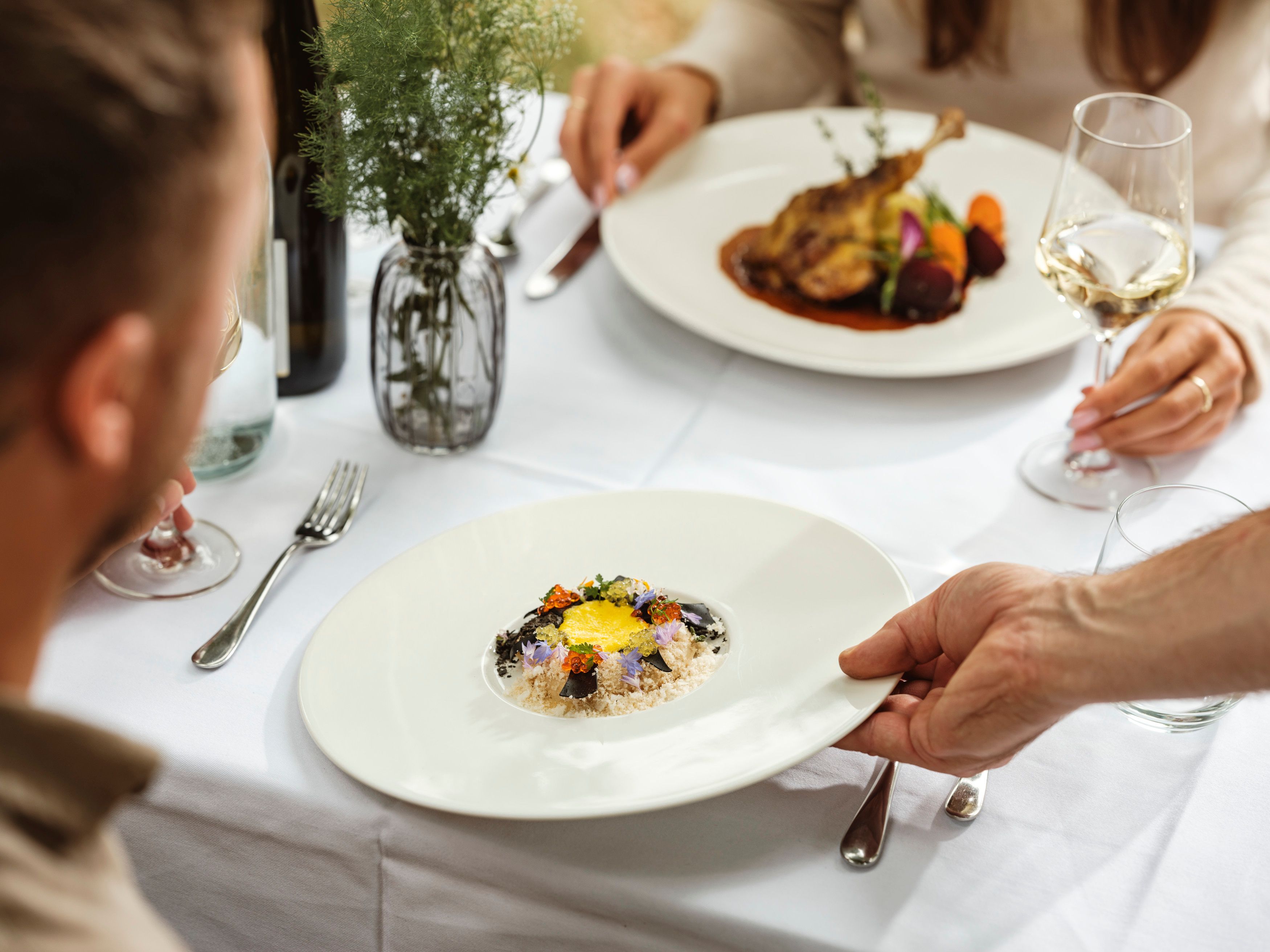 Ein elegantes Restaurantessen mit zwei Personen, die an einem Tisch sitzen. Ein Teller mit einem kunstvoll angerichteten Gericht wird serviert.