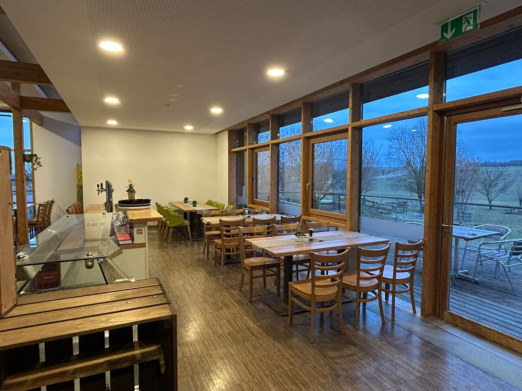 Innenansicht eines leeren Cafés mit Holzmöbeln und großen Fenstern mit Blick auf eine ländliche Landschaft.