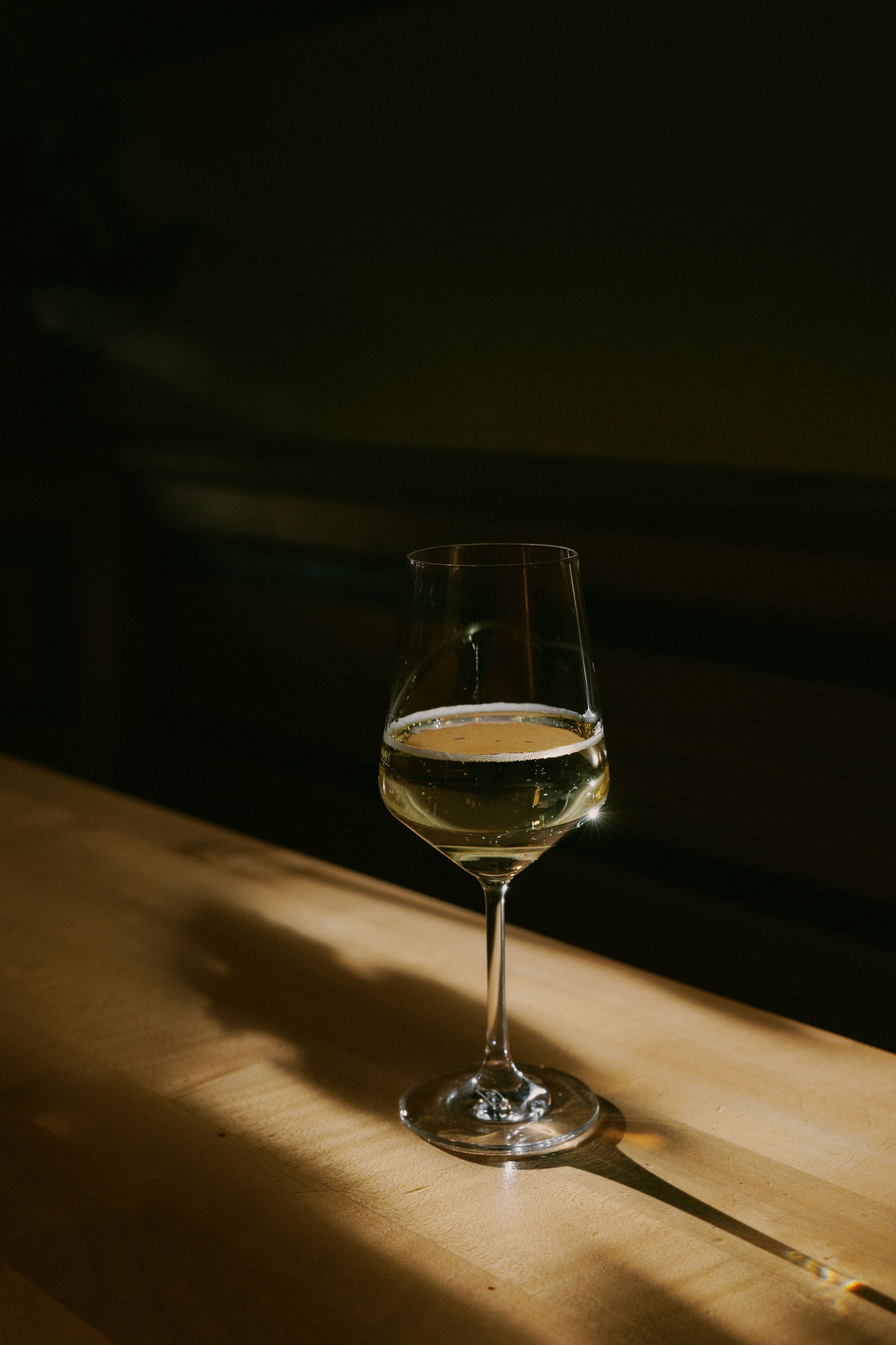 A wine glass with white wine stands on a wooden table, illuminated by a warm beam of light.
