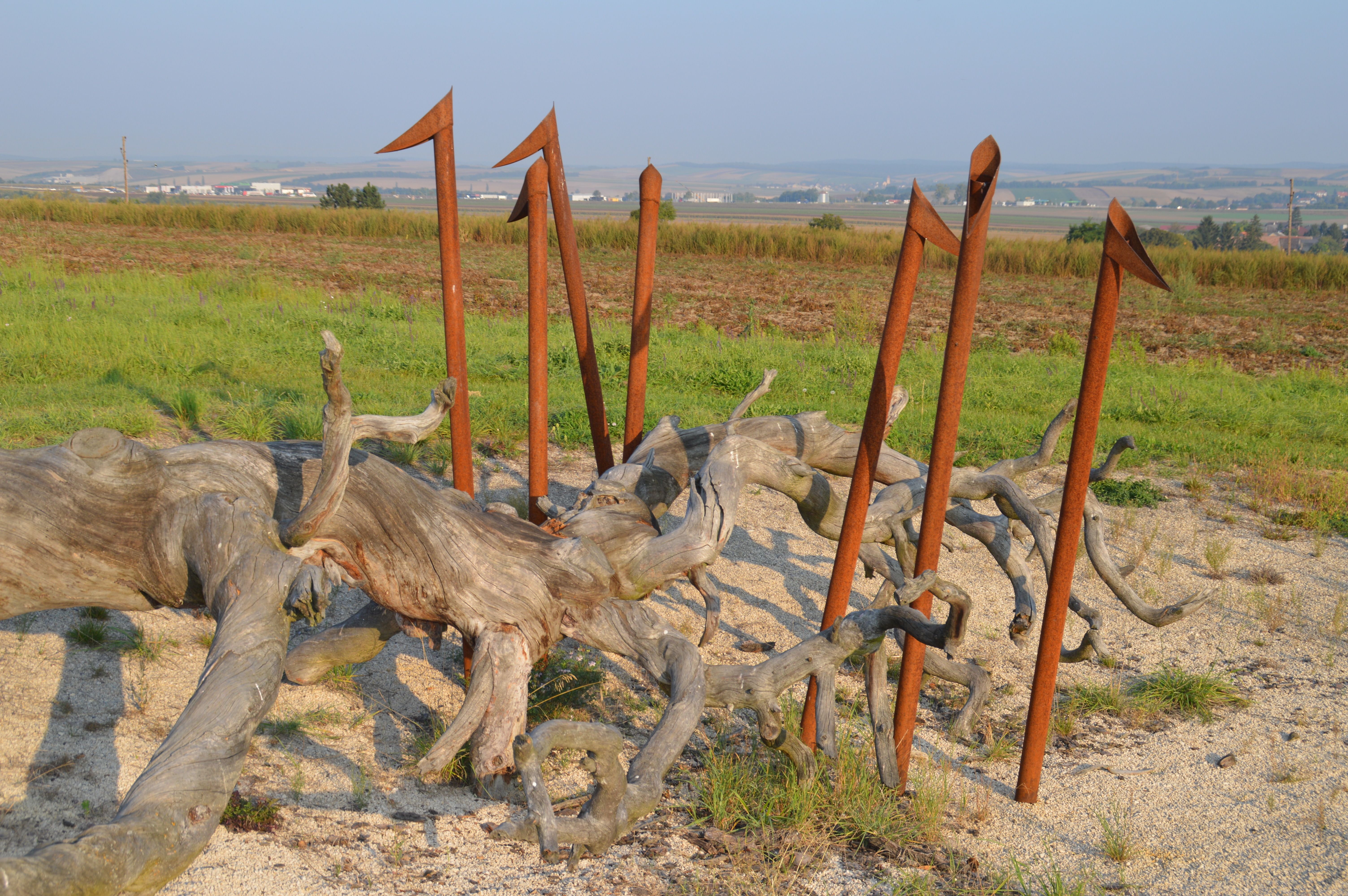 Kunstinstallation aus Holz und Metallstangen in einer ländlichen Landschaft.