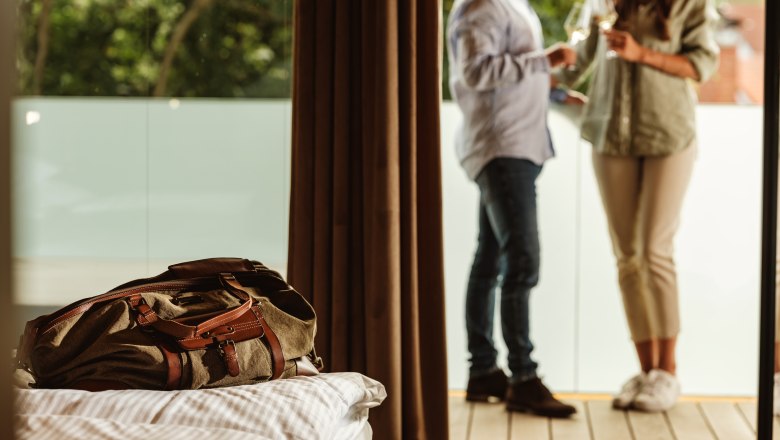 Herzlich Willkommen, © Weinviertel Tourismus GmbH / Michael Reidinger Ein Paar steht auf einem Balkon und stößt mit Gläsern an, im Vordergrund liegt eine Reisetasche auf einem Bett.