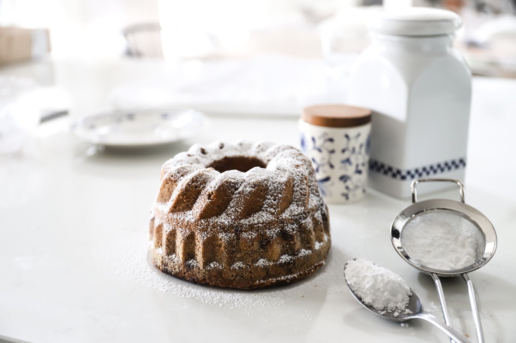 Ein Gugelhupf mit Puderzucker auf einem Tisch, daneben ein Sieb und ein Löffel mit Puderzucker.