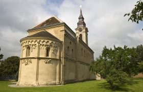 Romanische Kirche mit Verzierungen und Turm, umgeben von grüner Wiese und Bäumen.