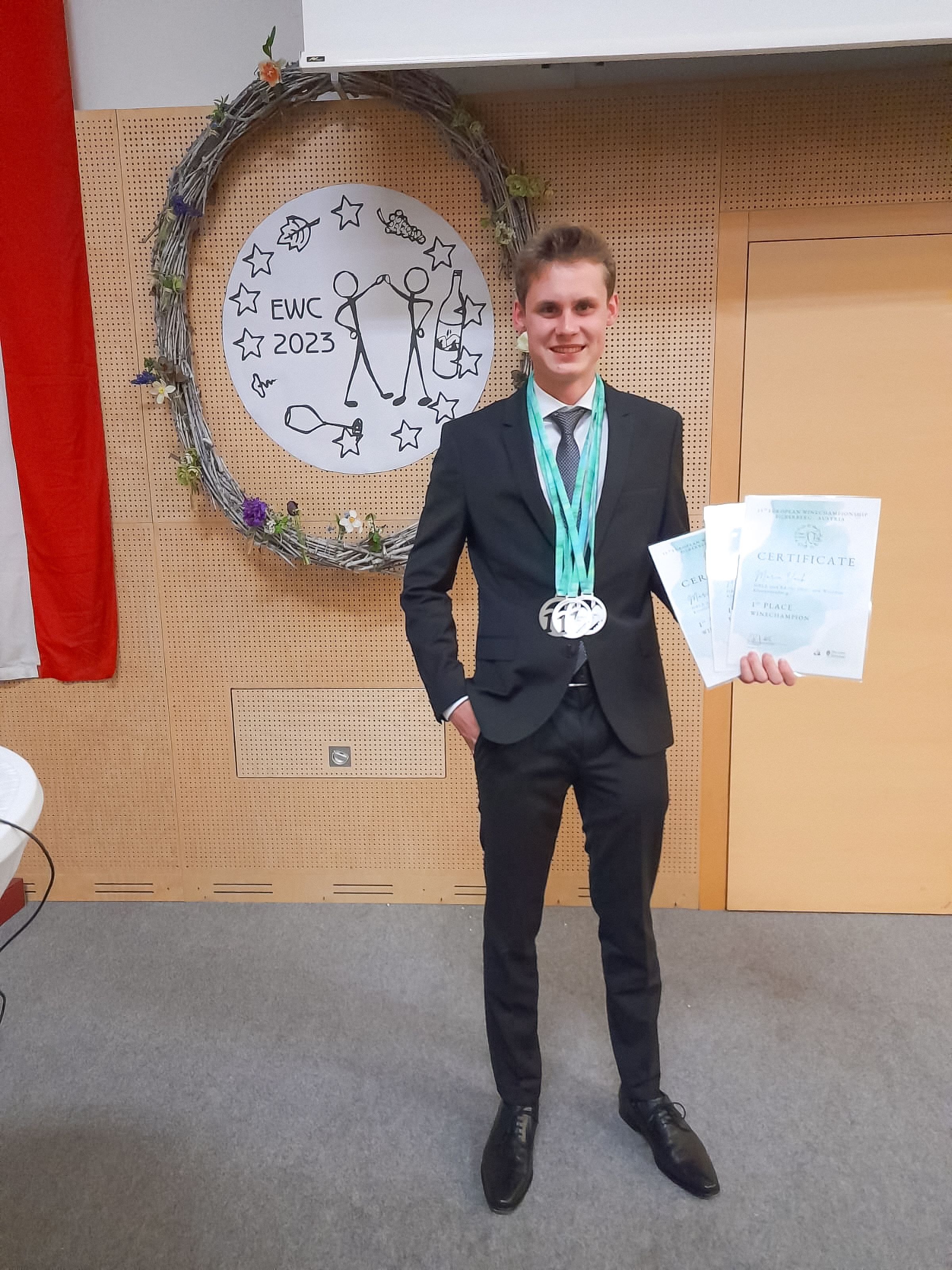 Ein junger Mann im Anzug steht vor einem dekorativen Kranz mit einem Zertifikat und Medaillen in der Hand.