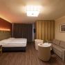 Modern furnished hotel room with double bed, sofa, two armchairs and a small table. Wooden floor and warm lighting.