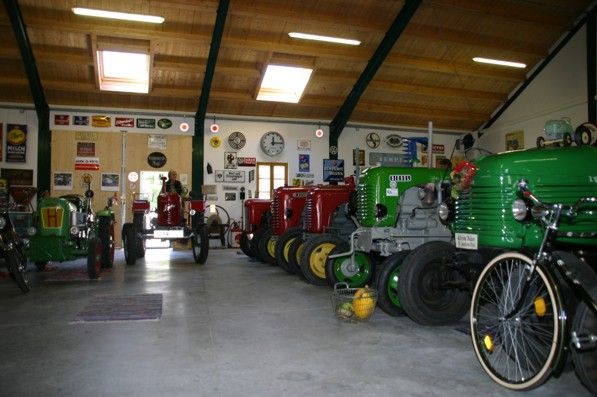 Innenansicht einer Scheune mit mehreren Oldtimer-Traktoren in verschiedenen Farben, umgeben von Retro-Werbeschildern an den Wänden.