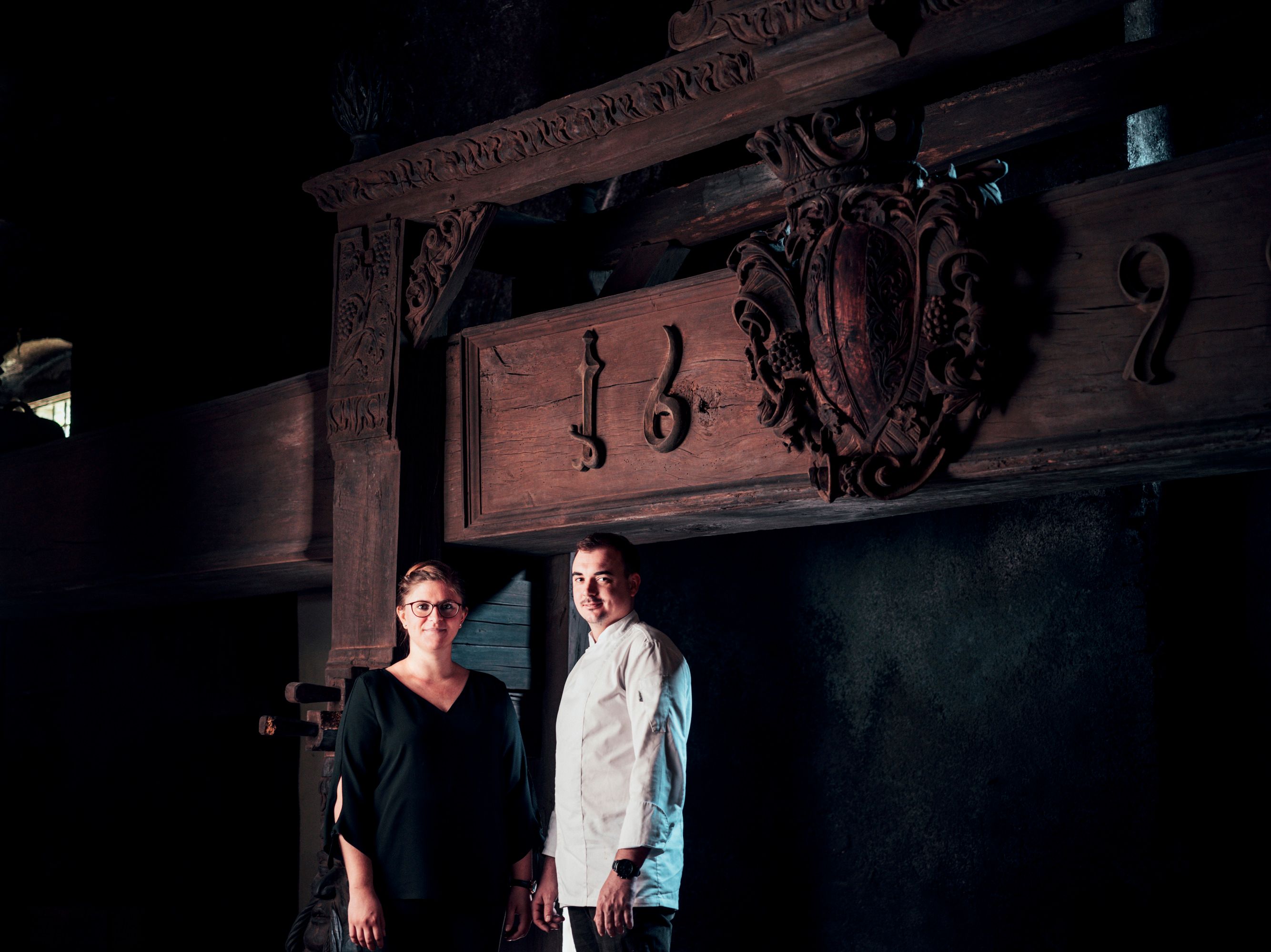 Zwei Personen stehen vor einem alten Holzbalken mit Schnitzereien und der Jahreszahl 1692.