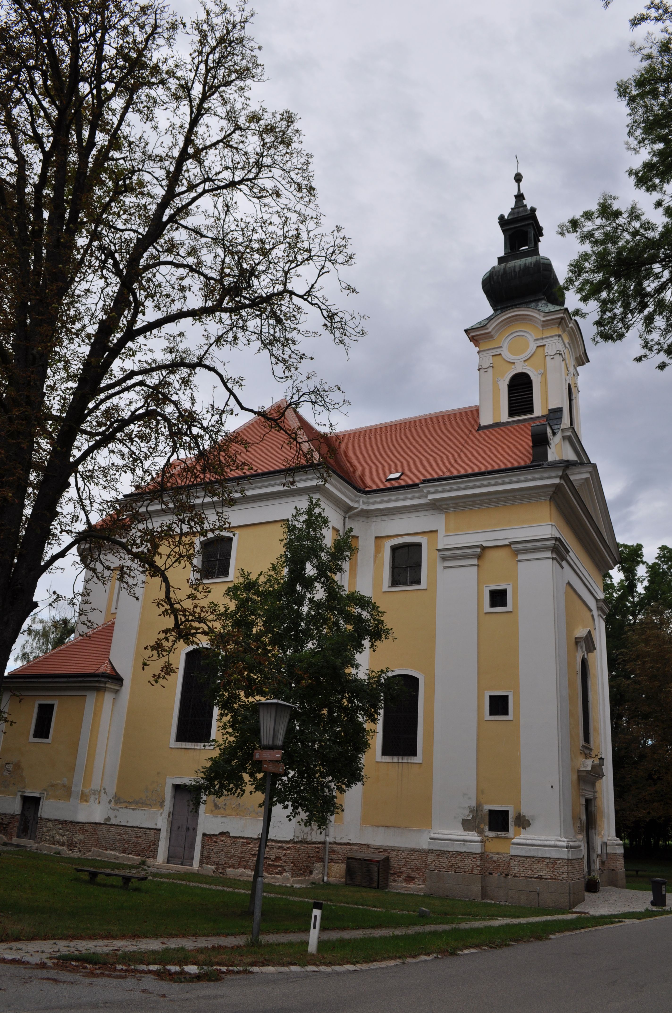 Žltý kostol s červenou strechou a vežou, obklopený stromami.