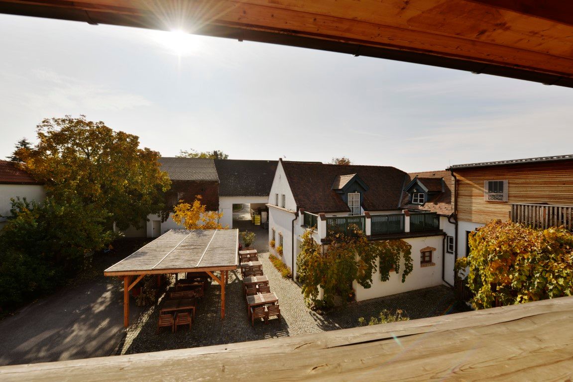 Hof eines Weinguts mit Tischen und Stühlen unter einem Holzpavillon, umgeben von Gebäuden und herbstlichem Laub.