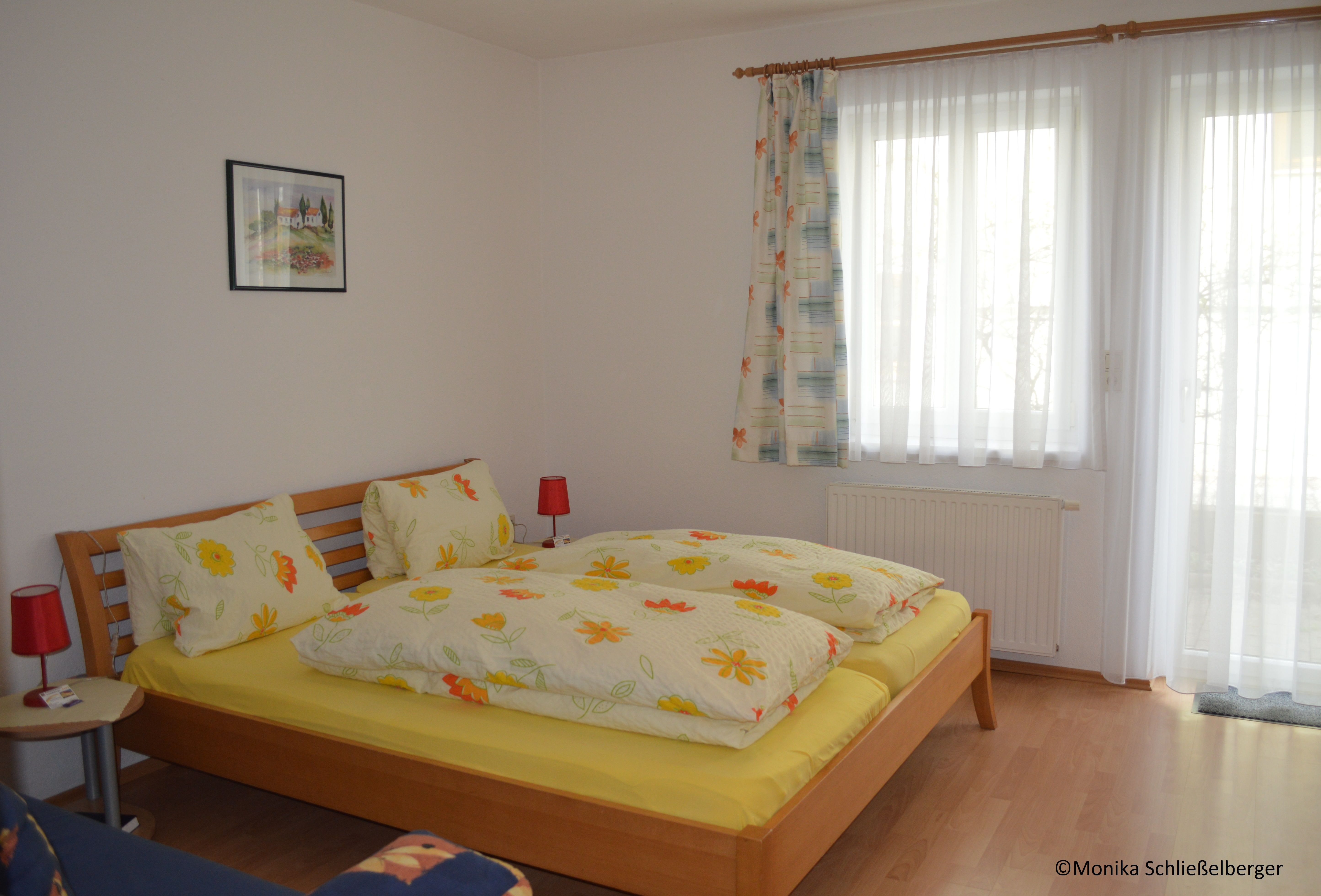 Ein gemütliches Doppelzimmer mit einem Holzbett, bunter Bettwäsche und zwei roten Nachttischlampen. Ein Fenster mit Vorhängen ist im Hintergrund.