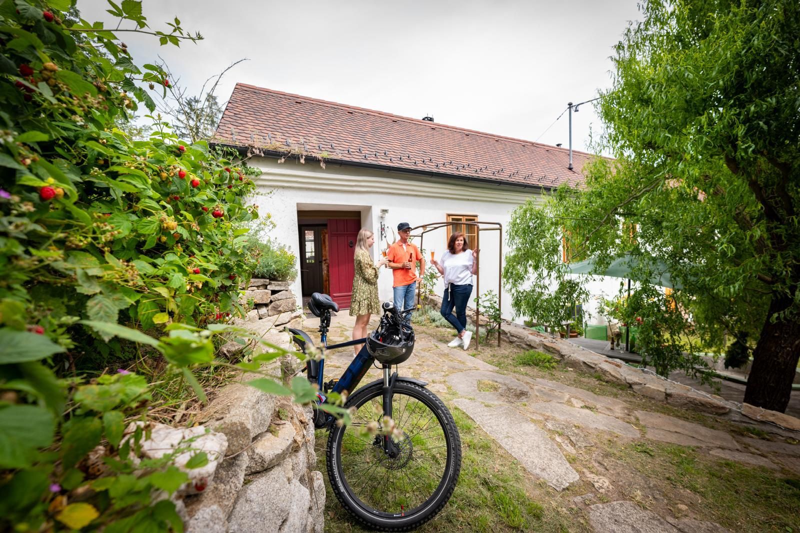 Drei Personen stehen vor einem weißen Haus mit rotem Dach, ein Fahrrad im Vordergrund.