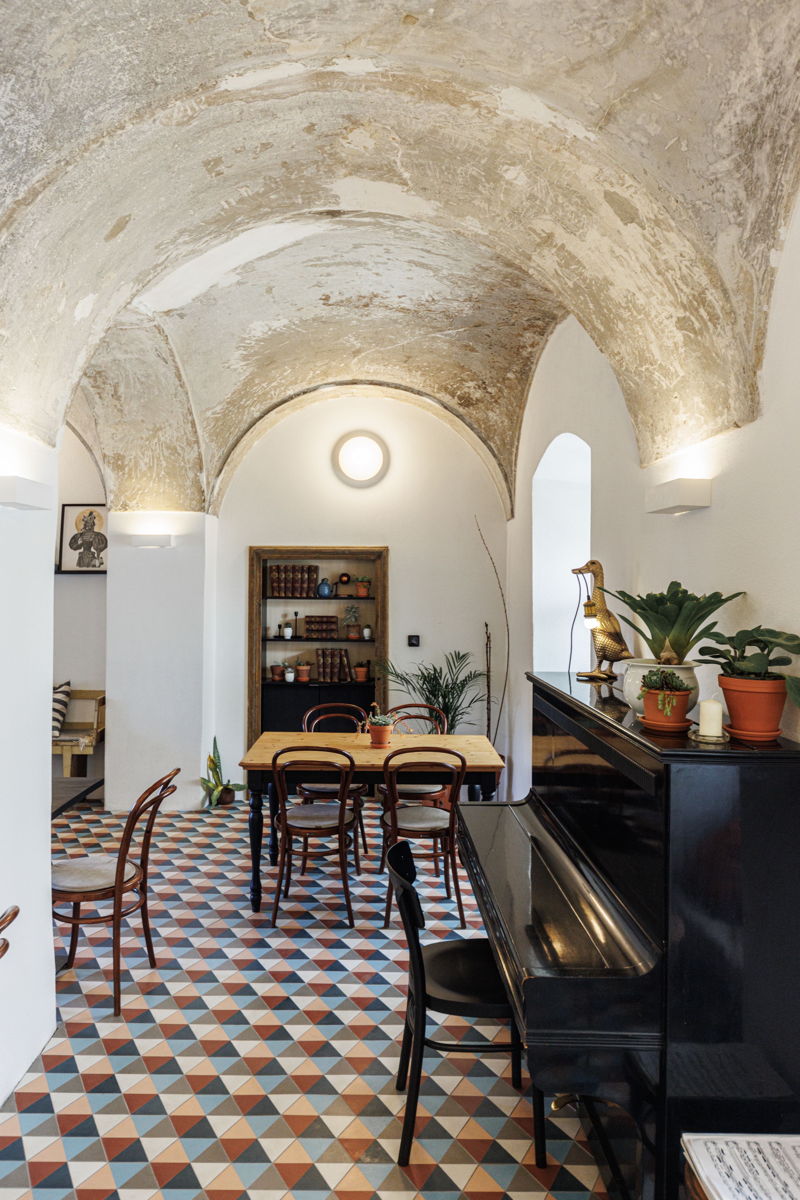 Ein stilvolles Café mit gewölbter Decke, buntem Fliesenboden, Holztisch und Stühlen. Ein Klavier steht rechts, Pflanzen und Dekorationen sind verteilt.