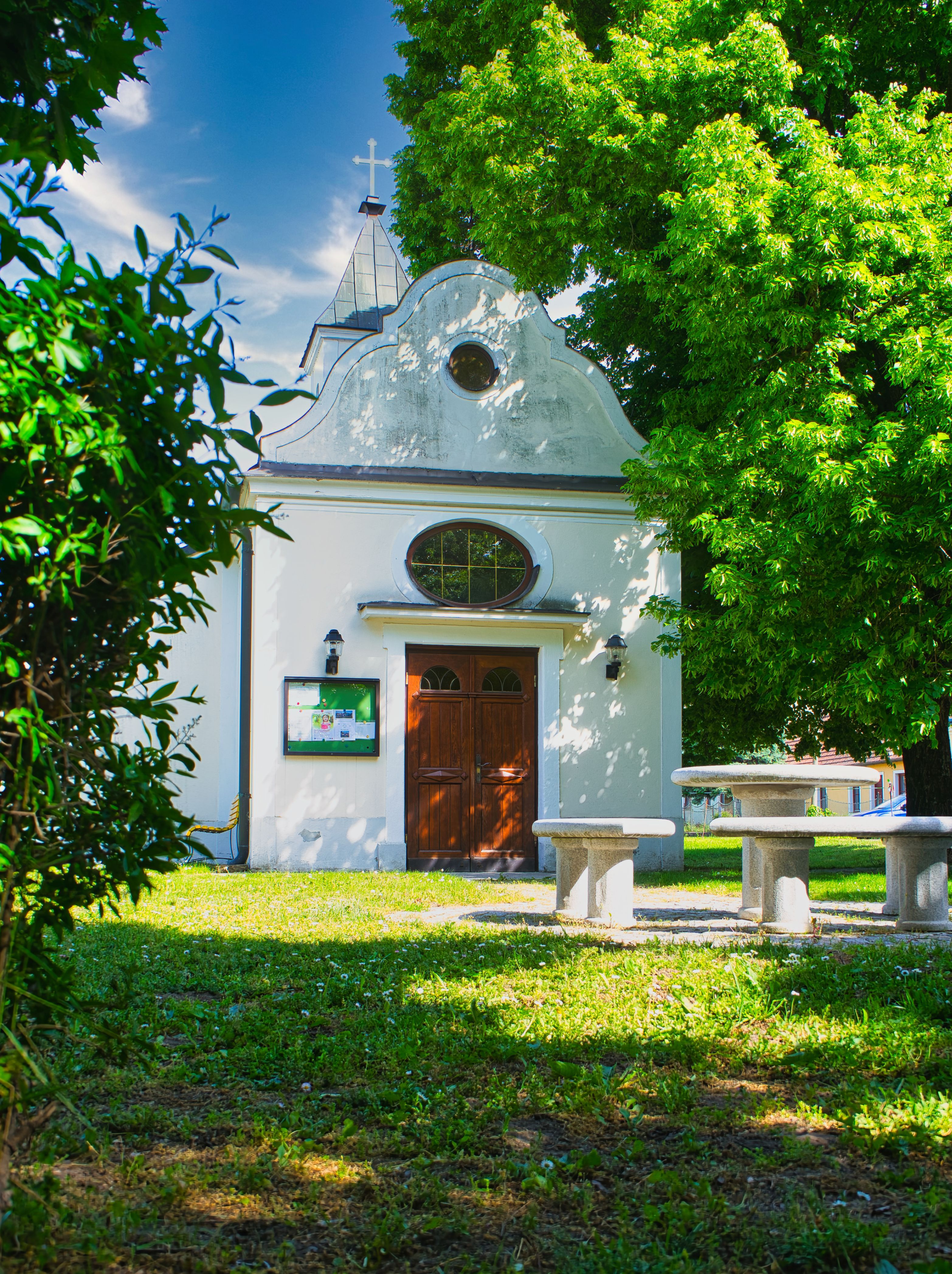Kleine Kapelle in der Gemeinde Großhofen mit Kreuz auf dem Dach, umgeben von Bäumen und Sitzbänken im Grünen.