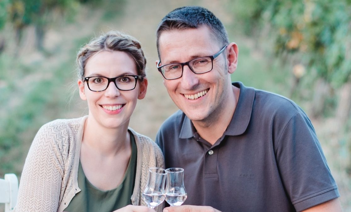 A smiling couple with glasses toast with shot glasses, blurred nature in the background.