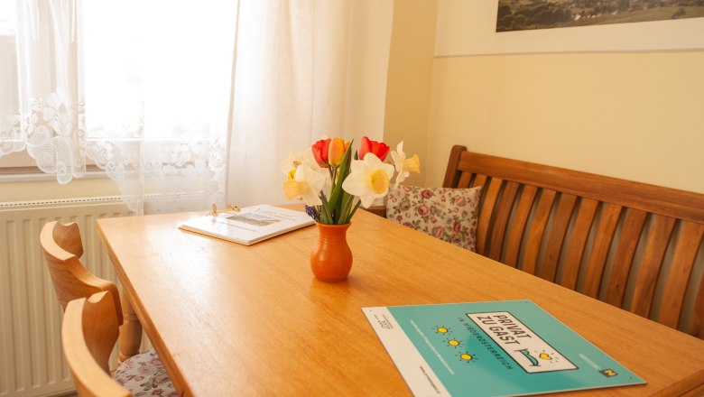 Ein gemütlicher Essbereich mit Holztisch, Blumenstrauß in Vase und Broschüren. Helle Vorhänge und ein Bild an der Wand.