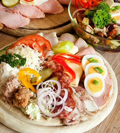 Various types of sausage and cheese on a wooden board, garnished with vegetables and eggs, next to a salad in a glass bowl.