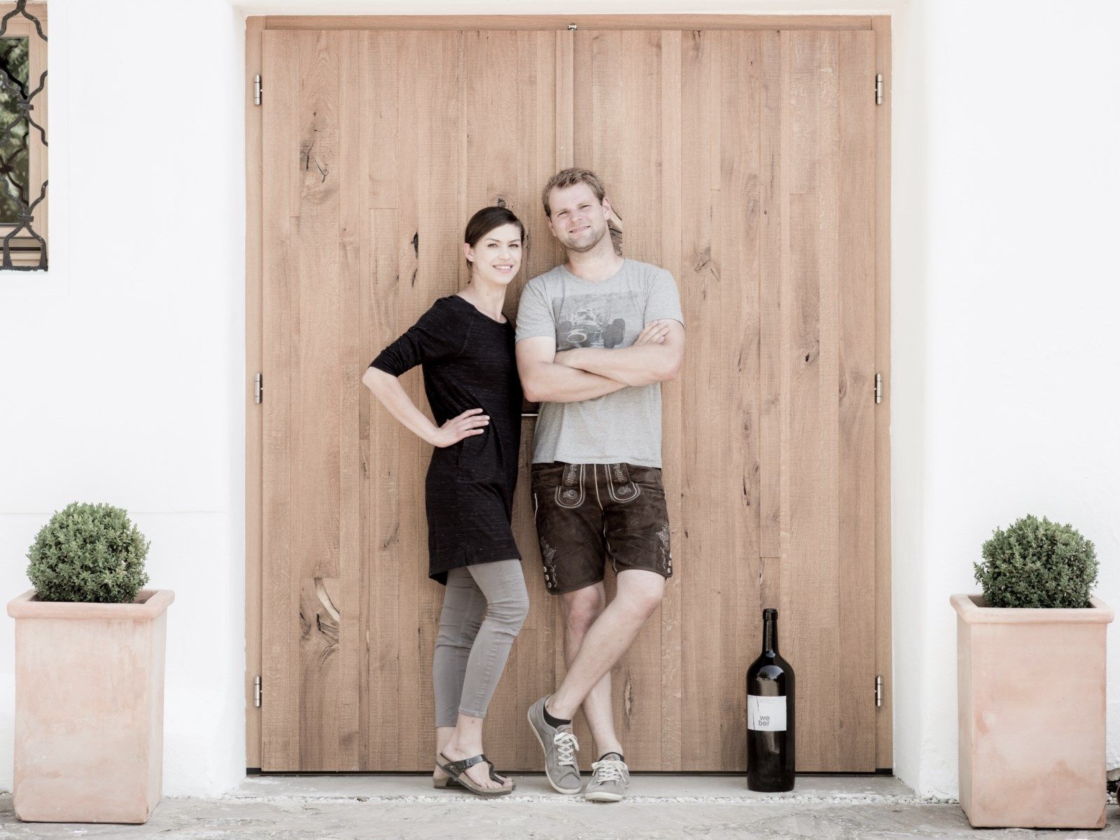 Zwei Personen stehen vor einer Holztür, daneben eine große Weinflasche und Pflanzen in Töpfen.