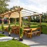 Holzpergola mit Bänken und Tischen im Freien, umgeben von Rasen und Bäumen.