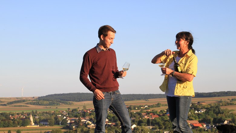 Two people are standing on a hill with wine glasses in their hands, in the background a rural landscape with wind turbines.