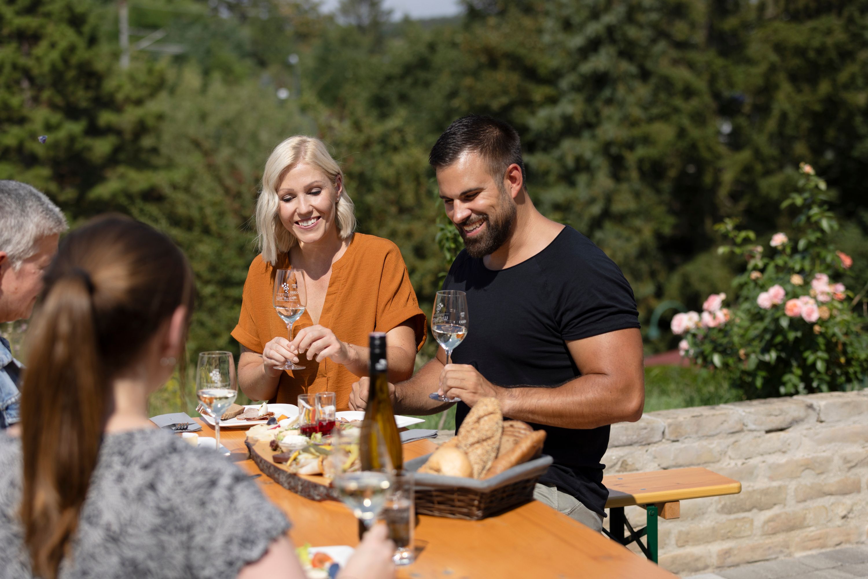 Gruppe von Menschen sitzt an einem Tisch im Freien und genießt Wein und Brot.