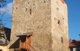 Ein alter Wehrturm mit hölzerner Treppe und Geländer, umgeben von Gebäuden, unter blauem Himmel.