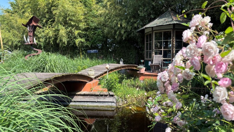 Ein idyllischer Garten mit Teich, Br&uuml;cke, Pavillon und bl&uuml;henden Rosen im Vordergrund.
