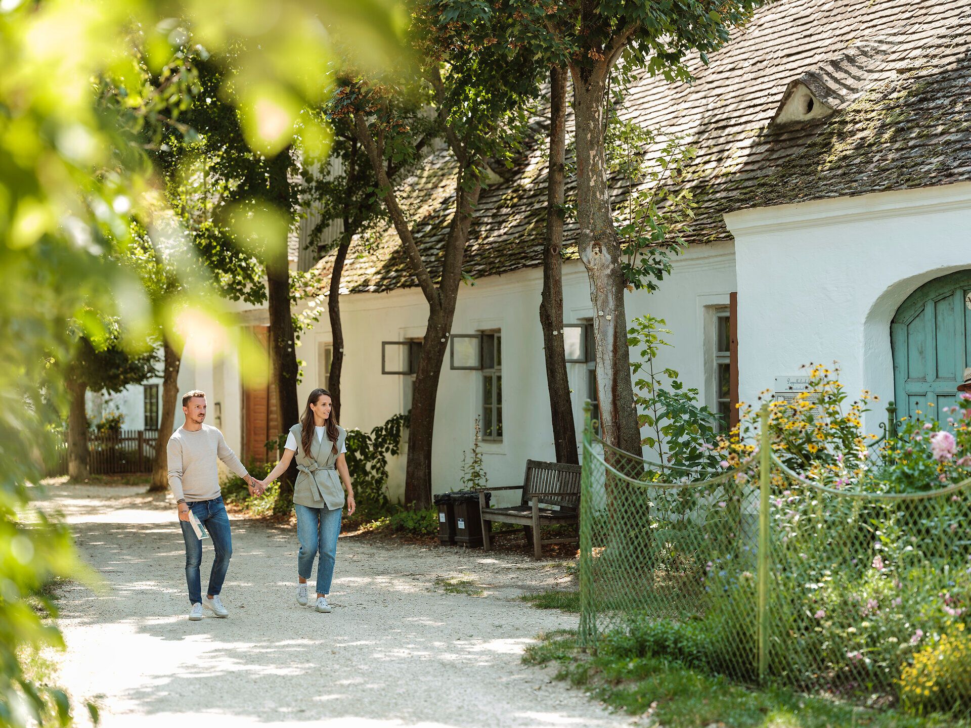 Inmitten der malerischen Kulisse des Weinviertler Museumsdorfes schlendern ein Paar Hand in Hand entlang der charmanten Dorfzeile. Die sanften Strahlen der Sonne tauchen die historischen Gebäude in warmes Licht und verleihen der Szenerie eine einladende Atmosphäre. Umgeben von üppigem Grün und blühenden Pflanzen wird der Sommer hier zu einem unvergesslichen Erlebnis.