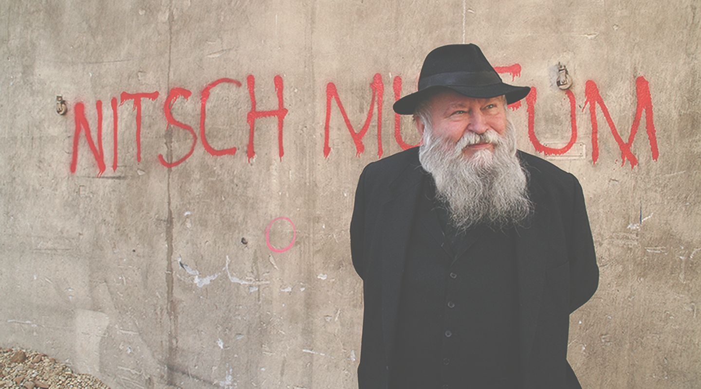Ein Mann mit Bart und Hut steht vor einer Wand mit der Aufschrift 'Nitsch Museum'.