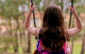 Ein Kind schaukelt im Freien, von hinten fotografiert, umgeben von B&auml;umen.