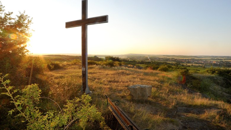 A cross on a hill at sunset with a view of a wide landscape and a village in the background.