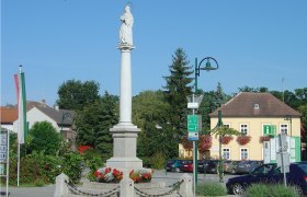 Statue auf einer S&auml;ule in einem Platz in Haugsdorf, umgeben von Geb&auml;uden und B&auml;umen.