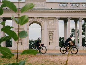 Radfahrer gleiten entspannt entlang der malerischen Landschaft, umgeben von &uuml;ppigem Gr&uuml;n und historischen S&auml;ulen. Die sanfte Brise und die beeindruckenden Ausblicke laden dazu ein, die Sch&ouml;nheit der Natur in vollen Z&uuml;gen zu genie&szlig;en.