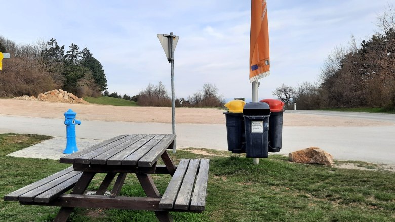 Picknicktisch und Mülleimer an einem Parkplatz mit Bäumen im Hintergrund.