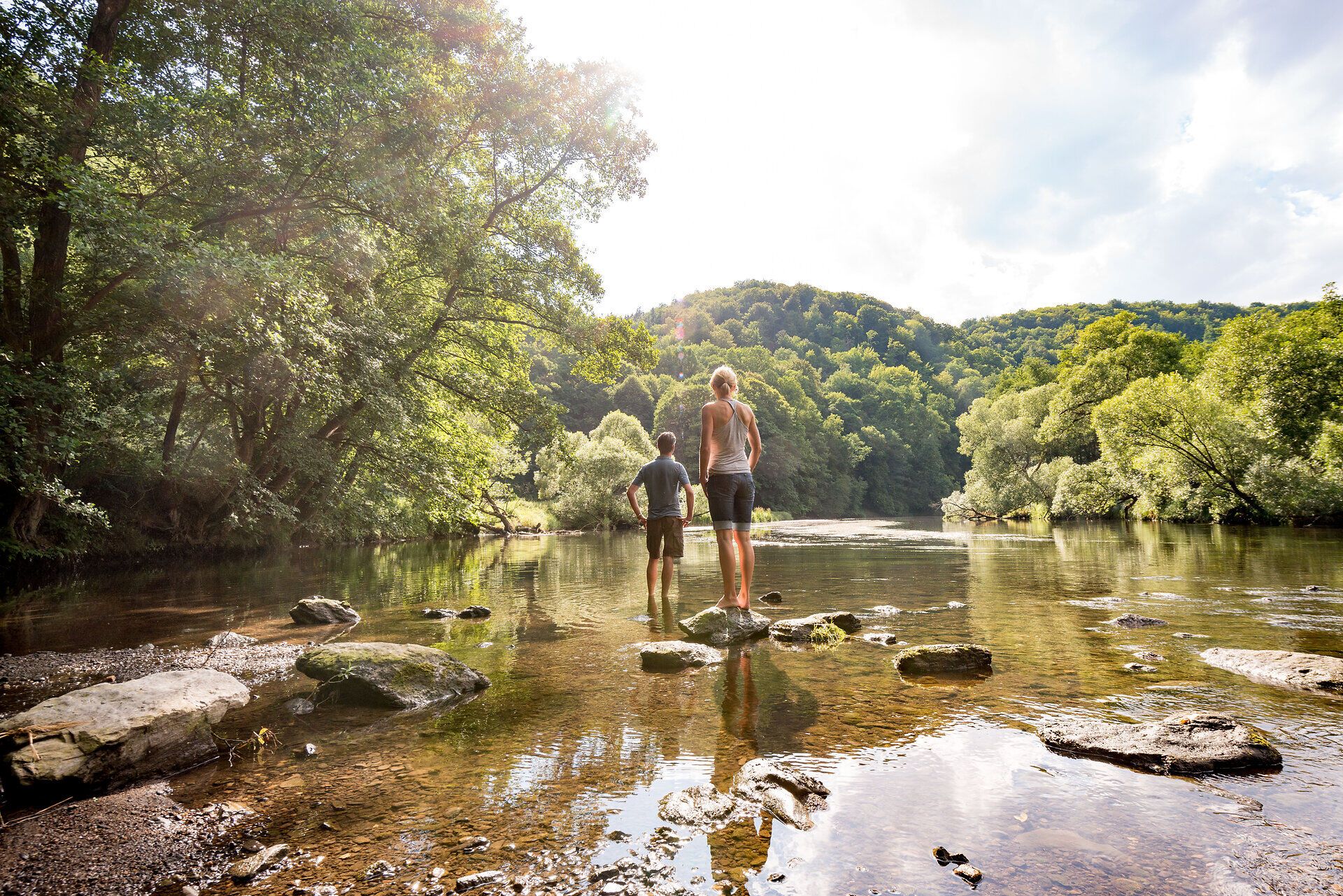 Ein sanfter Fluss schlängelt sich durch die malerische Landschaft, umgeben von üppigem Grün und majestätischen Bäumen. Zwei Wanderer stehen am Ufer und genießen die erfrischende Brise, während die Sonne durch die Wolken bricht und die Szenerie in goldenes Licht taucht.